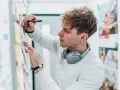 Ein junger Mann mit Kopfhörern am Hals und einem Stift in der Hand, der Ideen für sein Mediendesign Studium an einer Pinnwand sammelt.