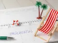 Kalenderblatt mit "Brückentag" markiert. Mini-Strandstuhl, Palmen und Rettungsring deuten auf Pfingstferien 2026 in Bayern hin.