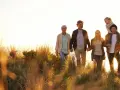 Eine Familie, die durch hohes Gras spaziert, zeigt eine schöne Generationen übersicht. Großeltern halten Händchen, Eltern gehen nebeneinander, ein Kind sitzt auf den Schultern des Vaters, ein weiterer Junge läuft daneben.