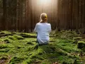 Frau sitzt im Wald auf moosbewachsenem Boden, umgeben von Bäumen. Sonnenstrahlen fallen durch das Blätterdach und erzeugen eine Atmosphäre der Katharsis.