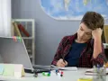 Ein Junge sitzt frustriert am Schreibtisch und macht seine hausaufgaben. Ein Laptop und bunte Stifte liegen bereit.
