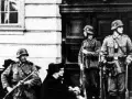 Soldaten der Gestapo stehen Wache. Ein Mann mit Gewehr im Vordergrund, zwei weitere im Hintergrund.