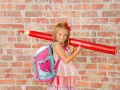 Ein Mädchen mit pinkem Rucksack und riesigem roten Stift vor Ziegelwand. Bereit für die Einschulung Berlin 2025!
