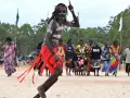 Ein junger Mann mit traditioneller Bemalung und rotem Lendenschurz tanzt. Im Hintergrund stehen Frauen und Männer, die die Zeremonie der Aborigines beobachten.