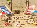 Strandkörbchen, Palmen und die Worte "Sommerferien" im Sand. Berlin wartet auf die Sommerferien 2026!