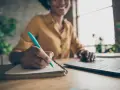 Lächelnde Frau schreibt Notizen in ein Notizbuch, während sie am Laptop arbeitet. Sie ist konzentriert, um ein Protokoll zu schreiben.
