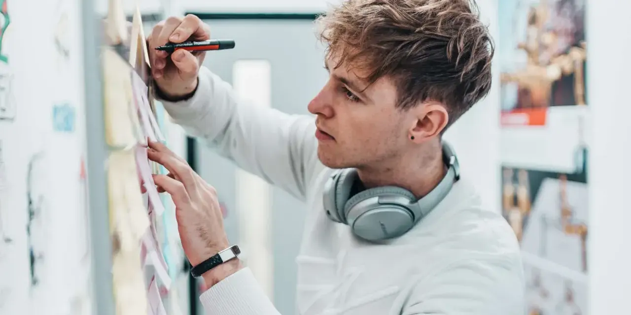 Ein junger Mann mit Kopfhörern am Hals und einem Stift in der Hand, der Ideen für sein Mediendesign Studium an einer Pinnwand sammelt.