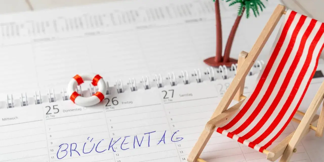 Kalenderblatt mit "Brückentag" markiert. Mini-Strandstuhl, Palmen und Rettungsring deuten auf Pfingstferien 2026 in Bayern hin.