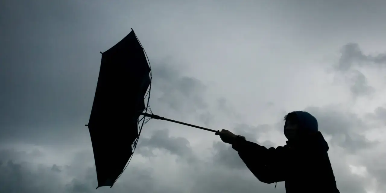 Silhouette einer Person mit einem aufgewehten Regenschirm vor dramatischem Himmel. Der **Orkan** kämpft gegen den Schirm.