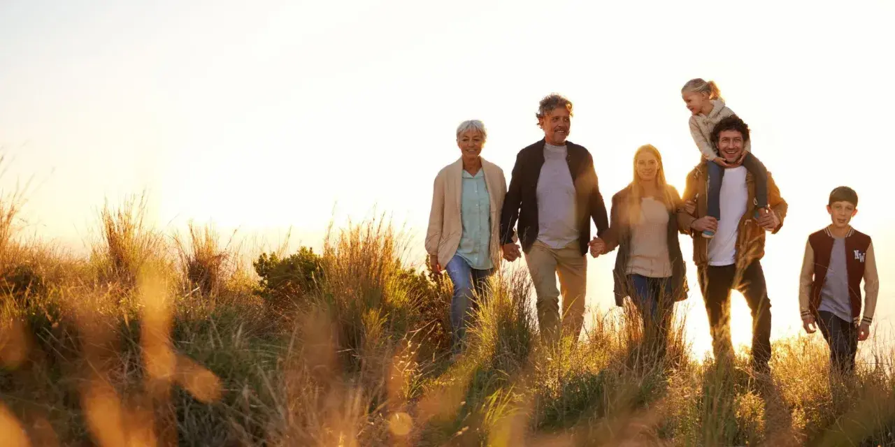 Eine Familie, die durch hohes Gras spaziert, zeigt eine schöne Generationen übersicht. Großeltern halten Händchen, Eltern gehen nebeneinander, ein Kind sitzt auf den Schultern des Vaters, ein weiterer Junge läuft daneben.
