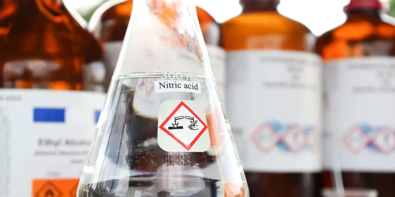 Ein Becherglas mit Salpetersäure (HNO3) und einem Gefahrensymbol. Im Hintergrund stehen weitere braune Glasflaschen mit Chemikalien.