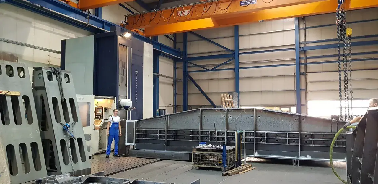 Ein junger Zerspanungsmechaniker in blauer Latzhose steht vor einer großen Soraluce-Maschine in einer Werkshalle. Daneben liegen massive Metallteile.