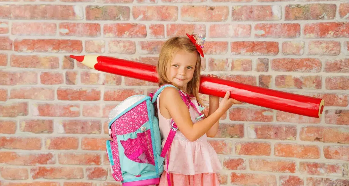 Ein Mädchen mit pinkem Rucksack und riesigem roten Stift vor Ziegelwand. Bereit für die Einschulung Berlin 2025!