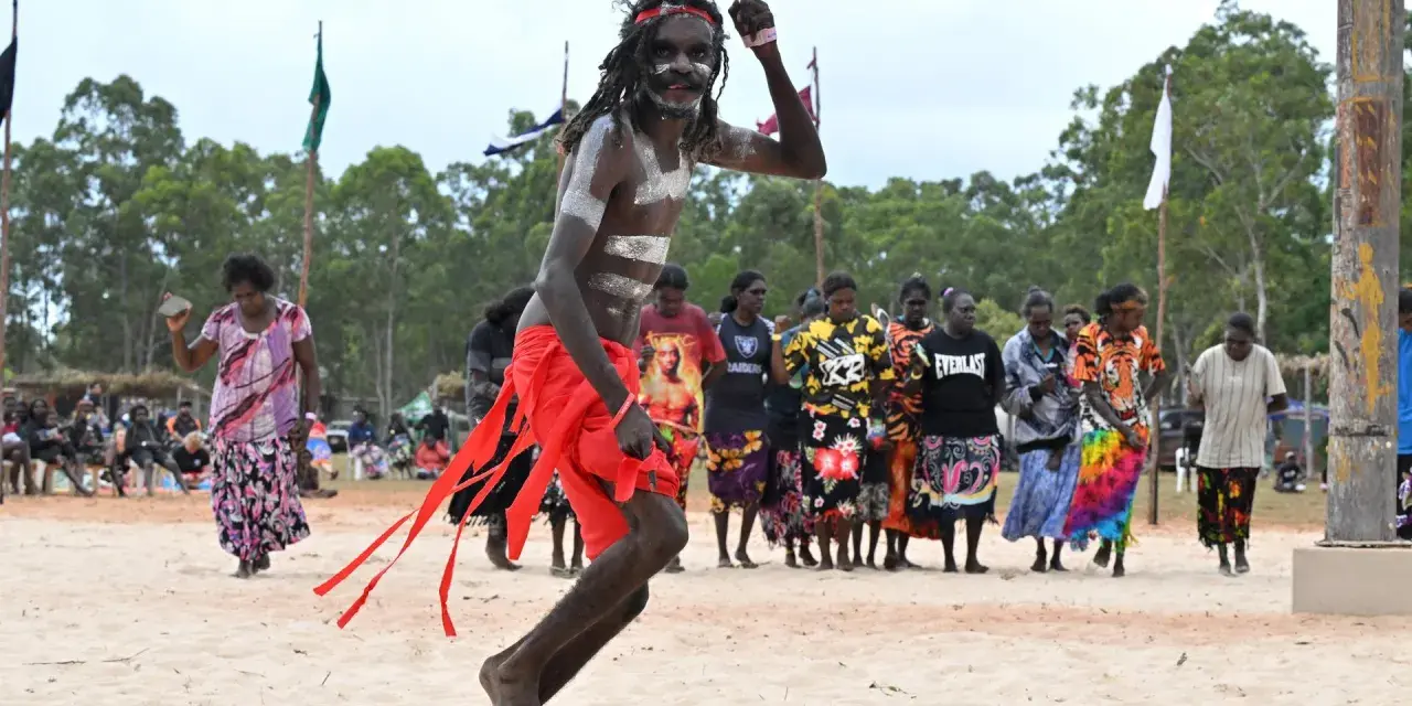 Ein junger Mann mit traditioneller Bemalung und rotem Lendenschurz tanzt. Im Hintergrund stehen Frauen und Männer, die die Zeremonie der Aborigines beobachten.