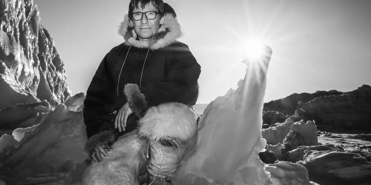Ein Mann in traditioneller Inuit-Kleidung sitzt in eisiger Landschaft. Die Sonne scheint hell.