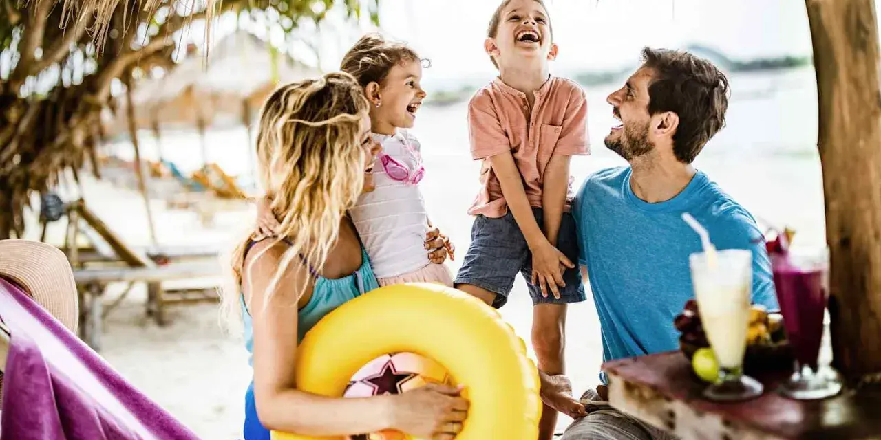 Lachende Familie am Strand mit gelbem Schwimmring und Getränken.