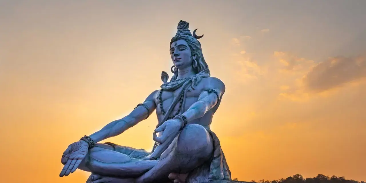 Eine riesige Statue von Shiva sitzt in meditativer Haltung vor einem warmen Sonnenuntergang.