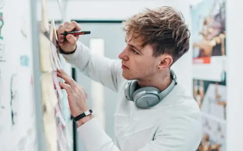 Ein junger Mann mit Kopfhörern am Hals und einem Stift in der Hand, der Ideen für sein Mediendesign Studium an einer Pinnwand sammelt.
