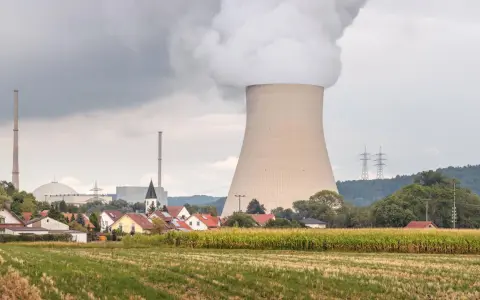 Dampf steigt aus einem Kühlturm eines Atomkraftwerks auf, daneben ein Dorf und ein Maisfeld.