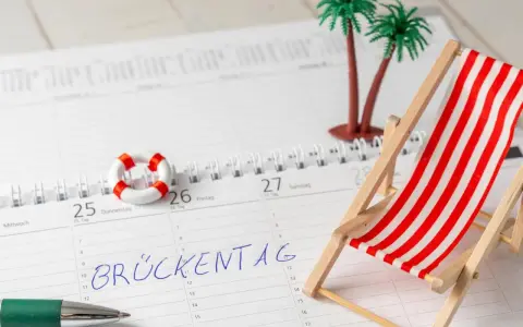 Kalenderblatt mit "Brückentag" markiert. Mini-Strandstuhl, Palmen und Rettungsring deuten auf Pfingstferien 2026 in Bayern hin.