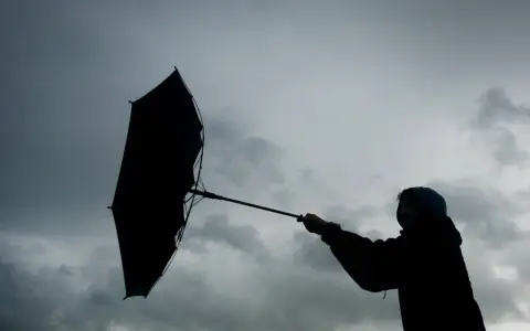 Silhouette einer Person mit einem aufgewehten Regenschirm vor dramatischem Himmel. Der **Orkan** kämpft gegen den Schirm.