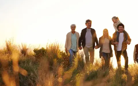 Eine Familie, die durch hohes Gras spaziert, zeigt eine schöne Generationen übersicht. Großeltern halten Händchen, Eltern gehen nebeneinander, ein Kind sitzt auf den Schultern des Vaters, ein weiterer Junge läuft daneben.