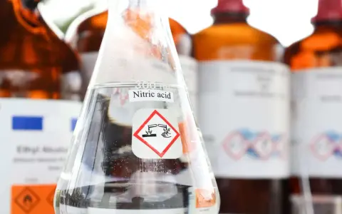 Ein Becherglas mit Salpetersäure (HNO3) und einem Gefahrensymbol. Im Hintergrund stehen weitere braune Glasflaschen mit Chemikalien.