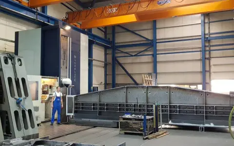 Ein junger Zerspanungsmechaniker in blauer Latzhose steht vor einer großen Soraluce-Maschine in einer Werkshalle. Daneben liegen massive Metallteile.