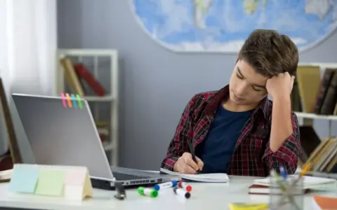 Ein Junge sitzt frustriert am Schreibtisch und macht seine hausaufgaben. Ein Laptop und bunte Stifte liegen bereit.