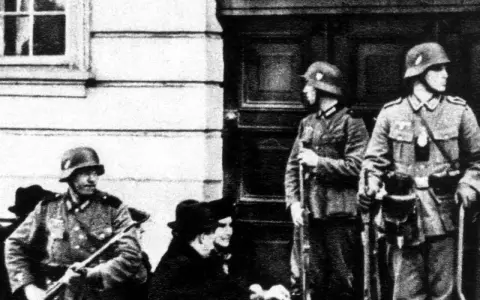Soldaten der Gestapo stehen Wache. Ein Mann mit Gewehr im Vordergrund, zwei weitere im Hintergrund.