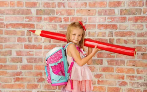 Ein Mädchen mit pinkem Rucksack und riesigem roten Stift vor Ziegelwand. Bereit für die Einschulung Berlin 2025!