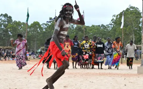 Ein junger Mann mit traditioneller Bemalung und rotem Lendenschurz tanzt. Im Hintergrund stehen Frauen und Männer, die die Zeremonie der Aborigines beobachten.