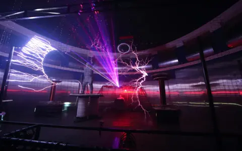 Spektakuläre Blitze zucken in einem **Faradayschen Käfig**, während ein Mann sicher darin steht. Laserstrahlen durchschneiden die Dunkelheit.