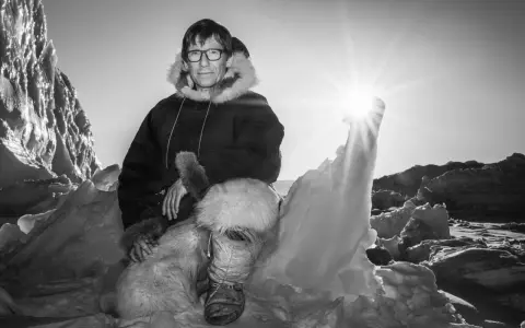 Ein Mann in traditioneller Inuit-Kleidung sitzt in eisiger Landschaft. Die Sonne scheint hell.
