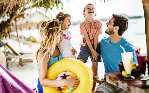 Lachende Familie am Strand mit gelbem Schwimmring und Getränken.