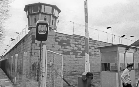 Wachturm und Grenzbefestigung der deutschen demokratischen Republik. Ein Wachmann steht neben einer Schranke und einem Kontrollhäuschen.
