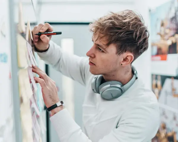 Ein junger Mann mit Kopfhörern am Hals und einem Stift in der Hand, der Ideen für sein Mediendesign Studium an einer Pinnwand sammelt.