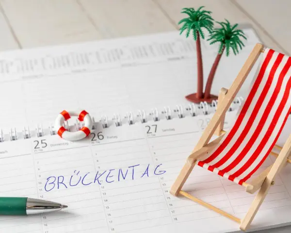 Kalenderblatt mit "Brückentag" markiert. Mini-Strandstuhl, Palmen und Rettungsring deuten auf Pfingstferien 2026 in Bayern hin.