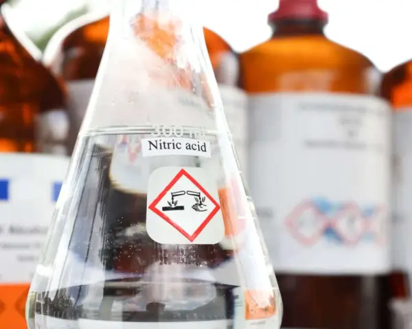 Ein Becherglas mit Salpetersäure (HNO3) und einem Gefahrensymbol. Im Hintergrund stehen weitere braune Glasflaschen mit Chemikalien.