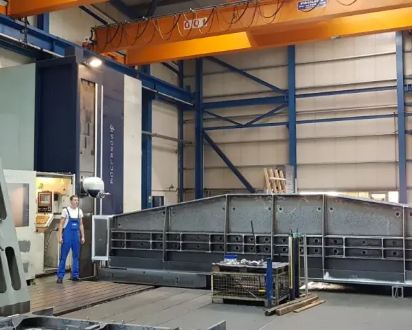 Ein junger Zerspanungsmechaniker in blauer Latzhose steht vor einer großen Soraluce-Maschine in einer Werkshalle. Daneben liegen massive Metallteile.