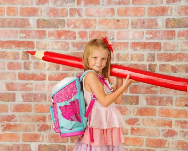 Ein Mädchen mit pinkem Rucksack und riesigem roten Stift vor Ziegelwand. Bereit für die Einschulung Berlin 2025!