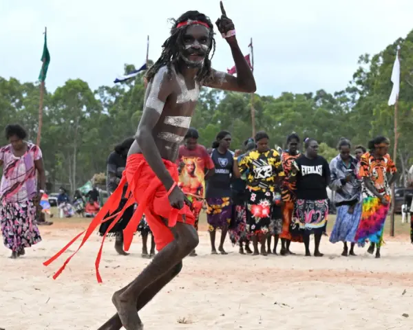 Ein junger Mann mit traditioneller Bemalung und rotem Lendenschurz tanzt. Im Hintergrund stehen Frauen und Männer, die die Zeremonie der Aborigines beobachten.