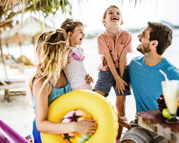 Lachende Familie am Strand mit gelbem Schwimmring und Getränken.