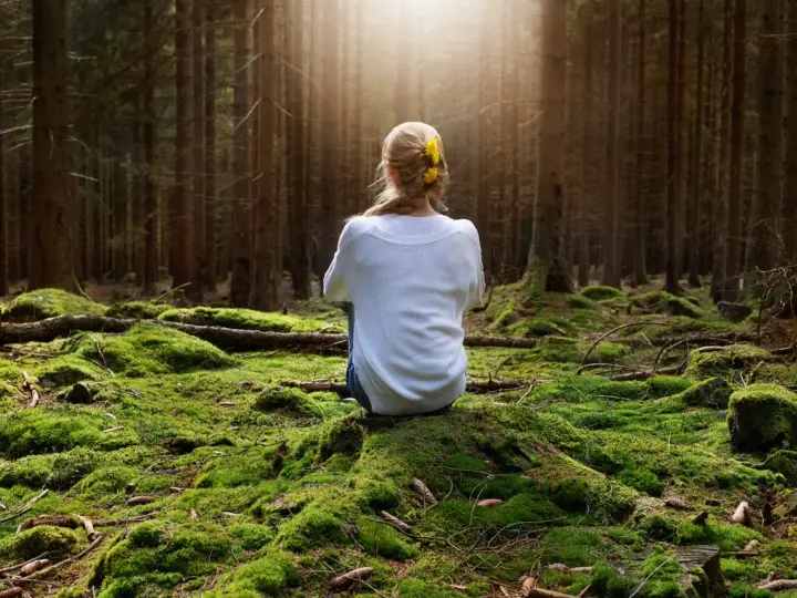 Frau sitzt im Wald auf moosbewachsenem Boden, umgeben von Bäumen. Sonnenstrahlen fallen durch das Blätterdach und erzeugen eine Atmosphäre der Katharsis.