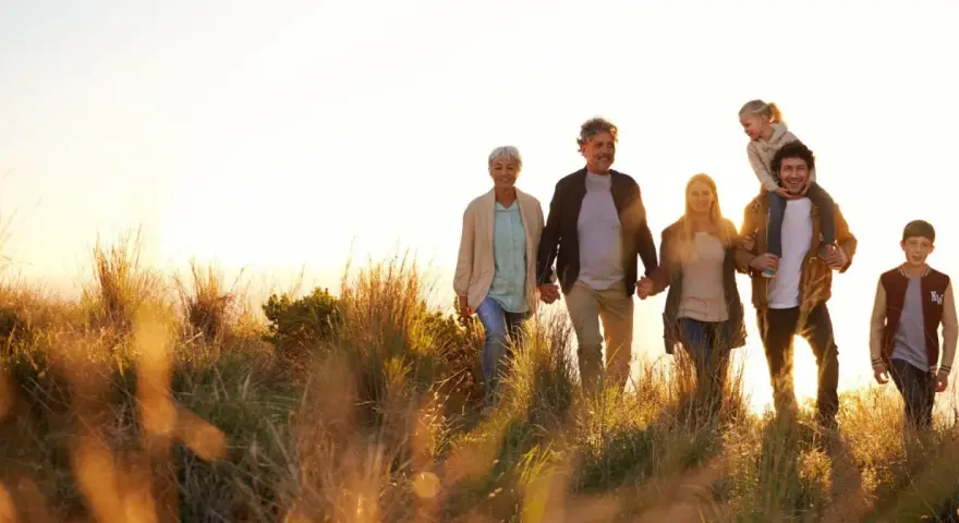 Eine Familie, die durch hohes Gras spaziert, zeigt eine schöne Generationen übersicht. Großeltern halten Händchen, Eltern gehen nebeneinander, ein Kind sitzt auf den Schultern des Vaters, ein weiterer Junge läuft daneben.