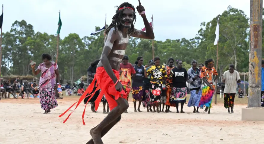 Ein junger Mann mit traditioneller Bemalung und rotem Lendenschurz tanzt. Im Hintergrund stehen Frauen und Männer, die die Zeremonie der Aborigines beobachten.