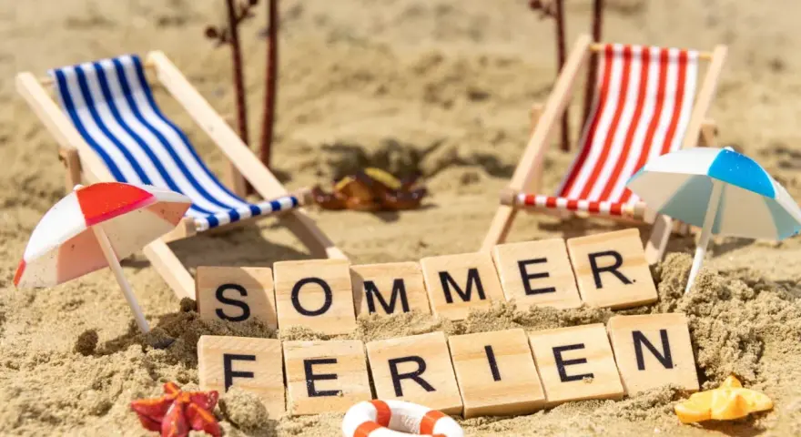 Strandkörbchen, Palmen und die Worte "Sommerferien" im Sand. Berlin wartet auf die Sommerferien 2026!