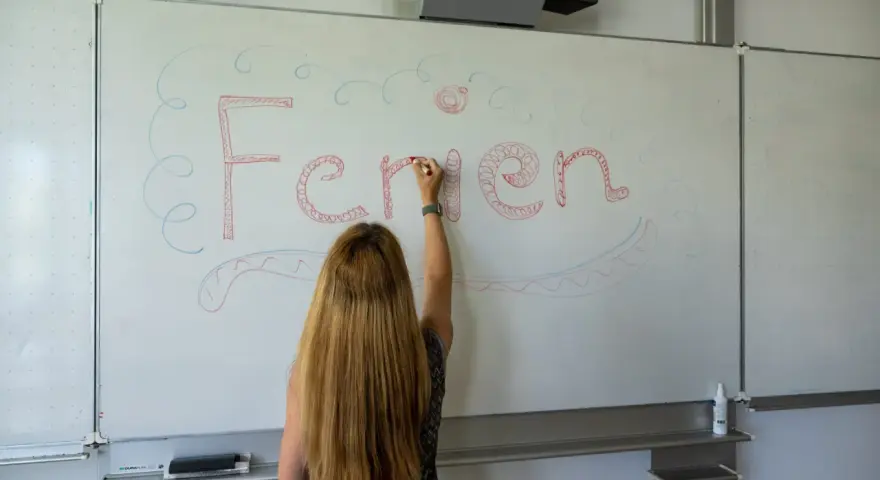 Frau schreibt "Ferien" an Tafel, Vorfreude auf Pfingstferien Rheinland-Pfalz.