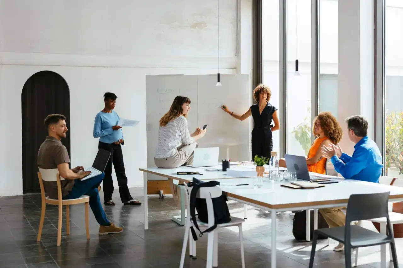Ein Team von Kollegen bespricht einen Marketingplan an einem Whiteboard. Eine Frau erklärt etwas, während andere zuhören und Notizen machen.