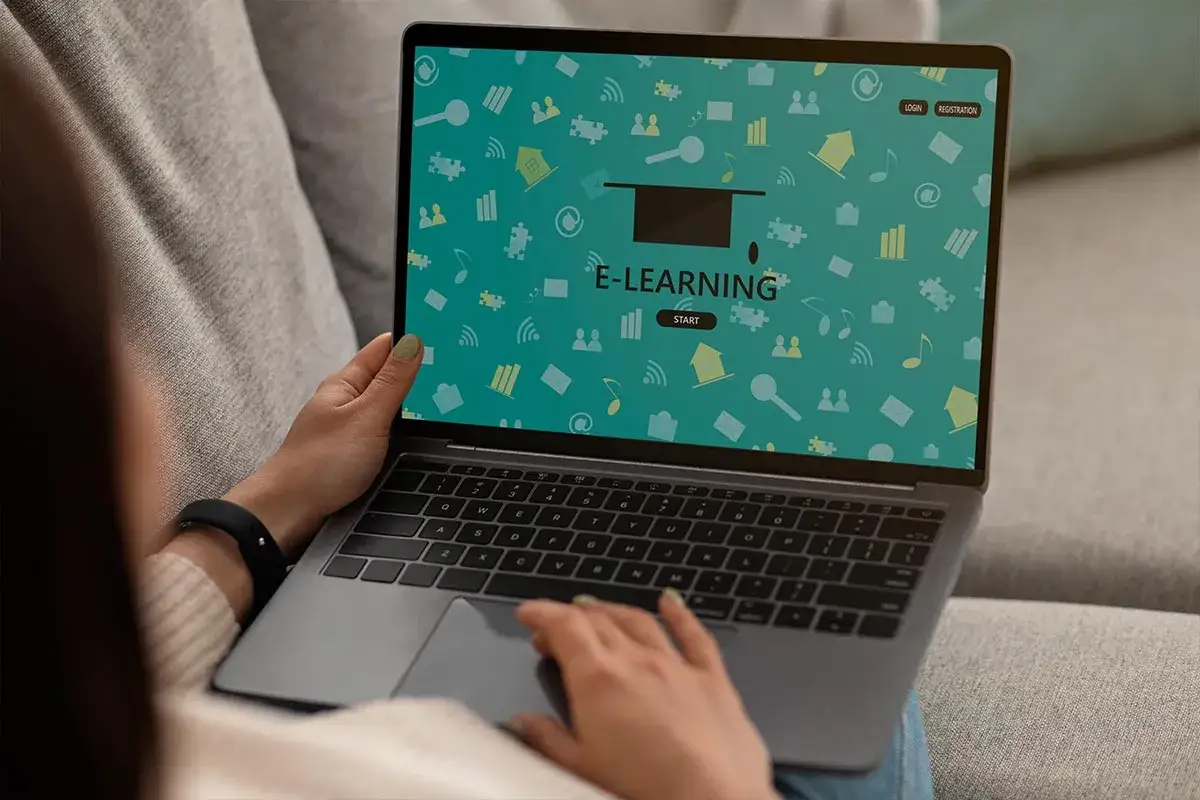 Person nutzt Laptop mit E-Learning-Interface. Bunte Icons wie Häuser, Puzzleteile und Noten zieren den Bildschirm.